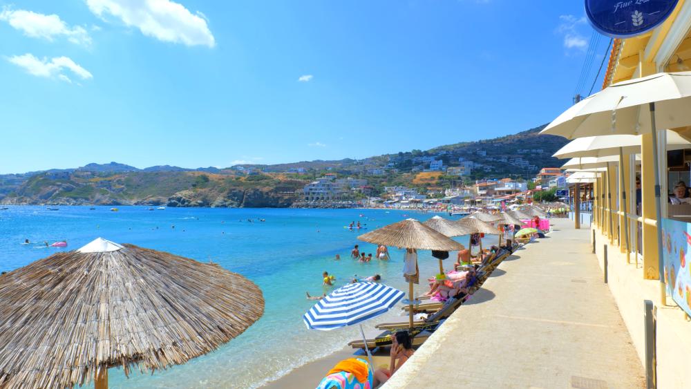 A vibrant beachfront in Agia Pelagia, Crete, Greece, featuring clear blue waters, sun umbrellas, and scenic hills in the background on a sunny day. | Smart Car Rental
