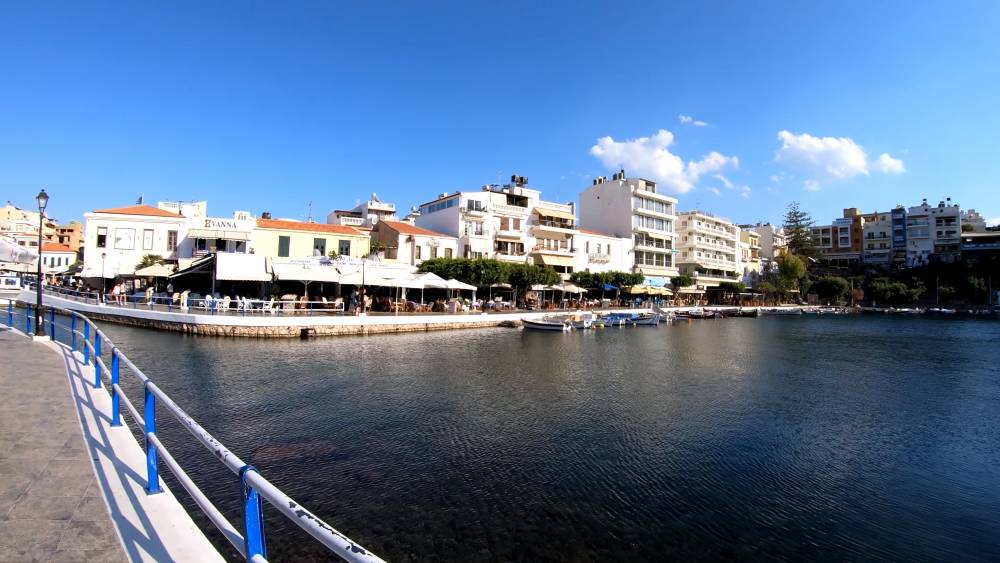 Een schilderachtig uitzicht op de waterkant in Agios Nikolaos, Kreta, met een rustige baai, boten en charmante gebouwen onder een heldere blauwe hemel. | SmartCarRental