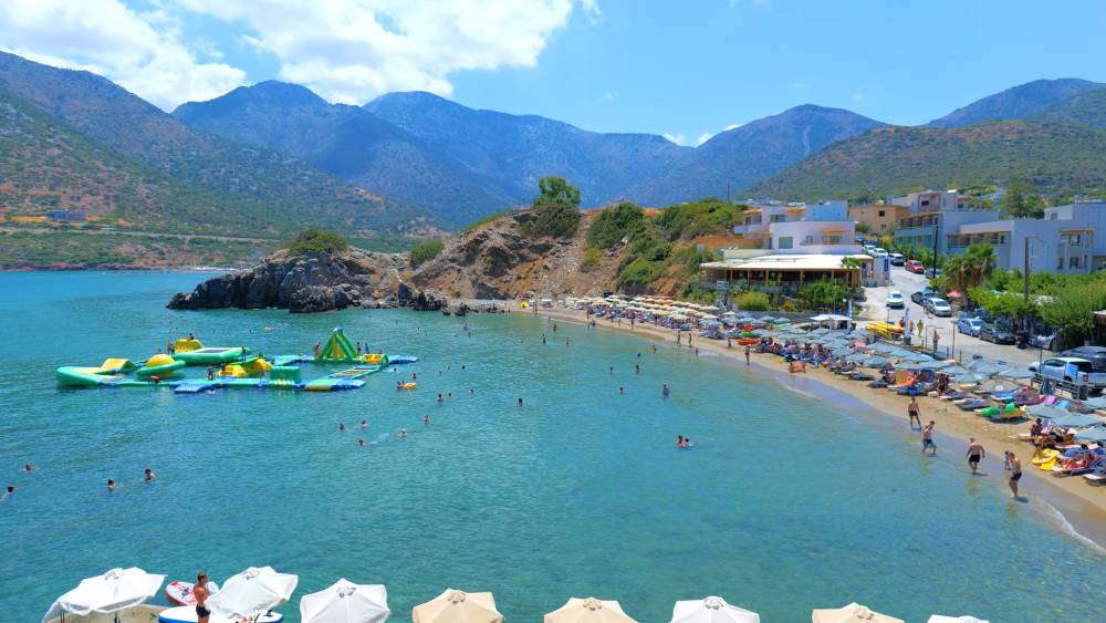 Vue de la plage à Bali, en Crète, avec une baie pittoresque aux eaux turquoise calmes, des personnes profitant des activités nautiques et du bronzage, et un arrière-plan de montagnes et de bâtiments côtiers. | Smart Car Rental