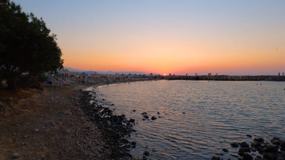 Zonsondergang uitzicht bij Gouves, Kreta, met een rustig strand met ligstoelen, een rotsachtige kustlijn en silhouetten van mensen die genieten van de serene sfeer aan zee. | Smart Car Rental