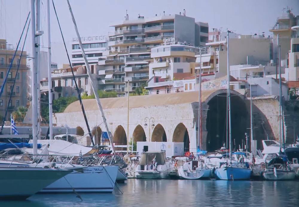 Uitzicht op boten aangemeerd in de haven van Heraklion op Kreta, met het Venetiaanse arsenaal en moderne gebouwen op de achtergrond, een combinatie van historische en eigentijdse architectuur. | Cheap Car Rental