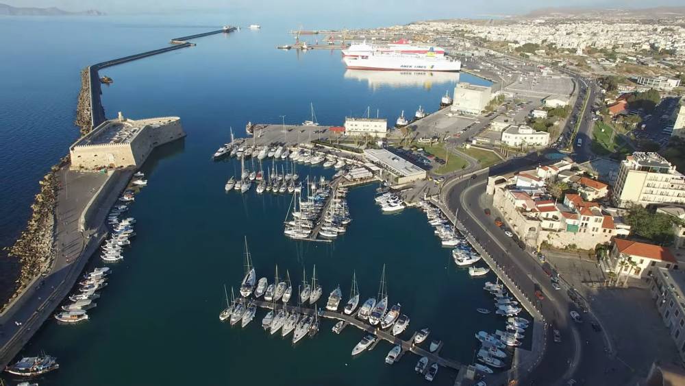 Drone uitzicht op de haven van Heraklion op Kreta, met talrijke boten aangemeerd in de jachthaven, het Venetiaanse fort en een grote veerboot op de achtergrond, met het stadsbeeld van Heraklion rondom de haven. | Smart Car Rental
