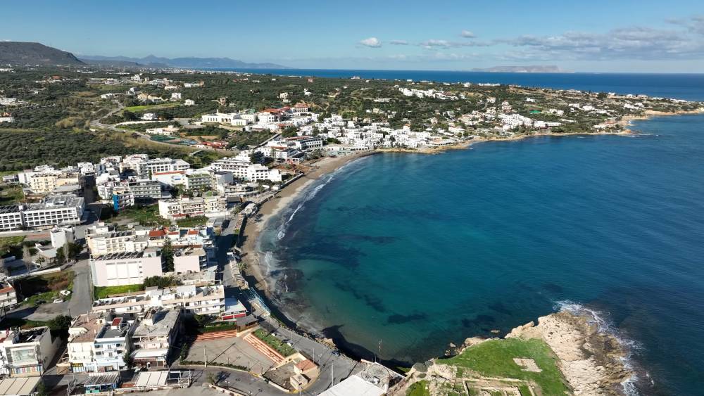 Widok z lotu ptaka na Hersonissos, ukazujący linię brzegową, budynki i przejrzystą błękitną wodę z perspektywy drona. | Cheap car rental
