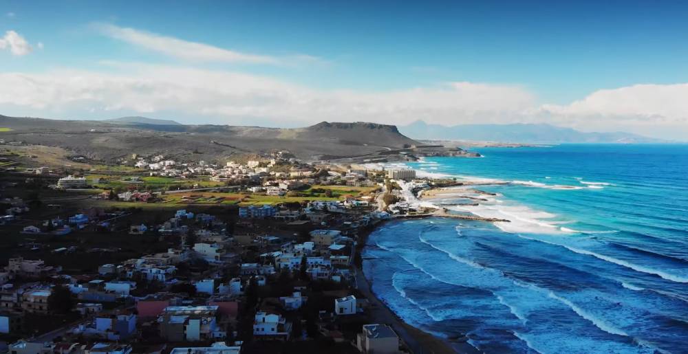 Luchtfoto van Kokkini Hani, Kreta, met een kustplaats met witte gebouwen, rollende golven langs de kustlijn en een bergachtig landschap onder een heldere blauwe hemel. | Cheap Car Rental