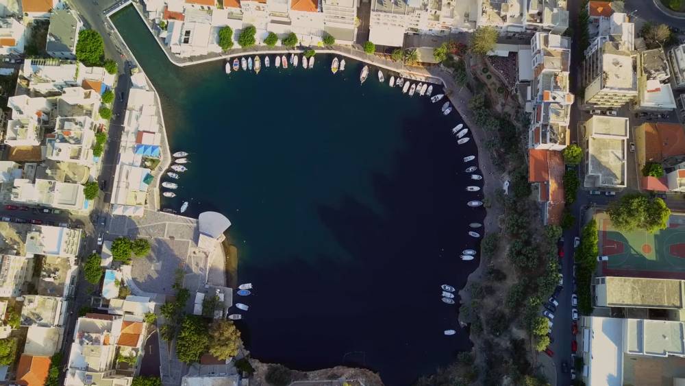 Een prachtige luchtfoto van Lake Voulismeni in Agios Nikolaos, Kreta, met het serene blauwe water omgeven door boten en gebouwen. | Cheap Car Rental