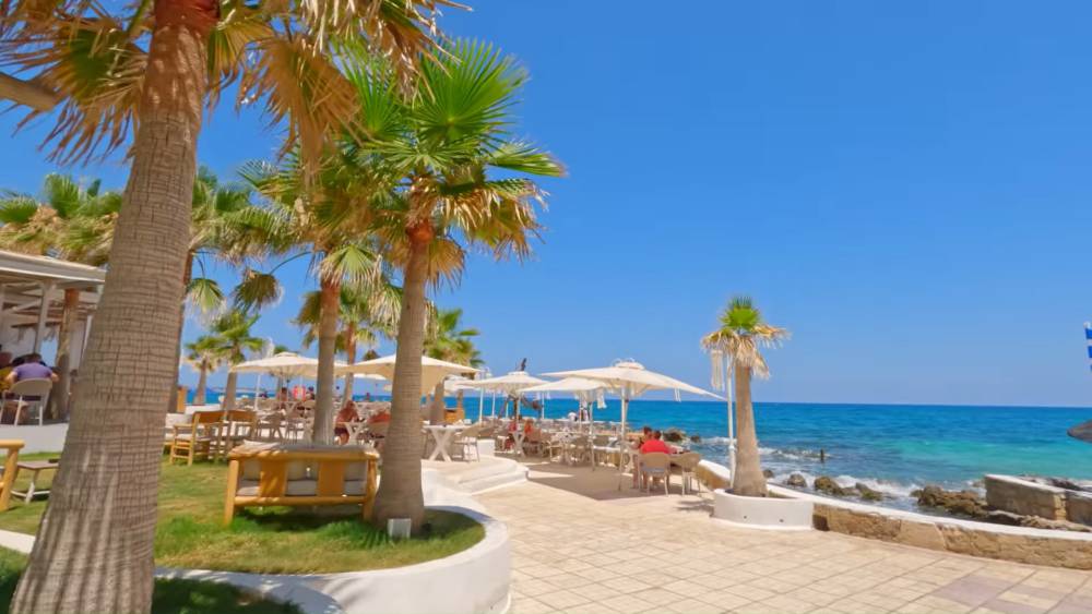 Een prachtig strandrestaurant in Malia, Kreta, Griekenland, met palmbomen, een buitenterras en het heldere blauwe water van de Middellandse Zee. | Cheap Car Rental