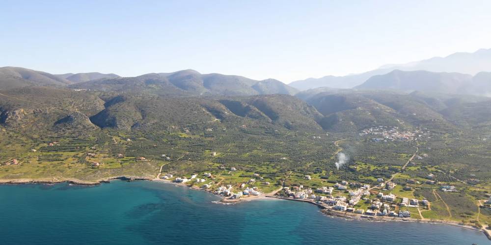 Een luchtfoto van Milatos, een kustdorp op Kreta, Griekenland, met groene heuvels, bergen en het heldere blauwe water van de Middellandse Zee. | Smart Car Rental