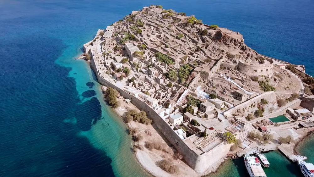 Een gedetailleerde luchtfoto van Spinalonga Eiland, Kreta, met oude ruïnes en vestingwerken omgeven door helderblauw water. | Cheap Car Rental